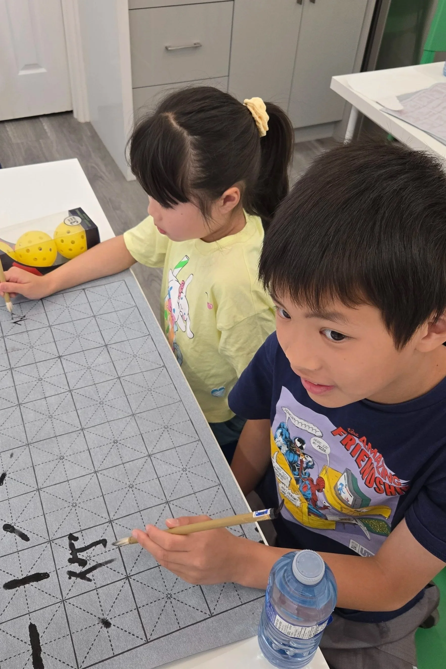 A boy and a girl sitting at a table with a grid mat, practicing calligraphy with brushes. The boy is writing, and there is a water bottle on the table. The girl is focused on writing, and there are yellow yellow dice and a paper towel on the table.