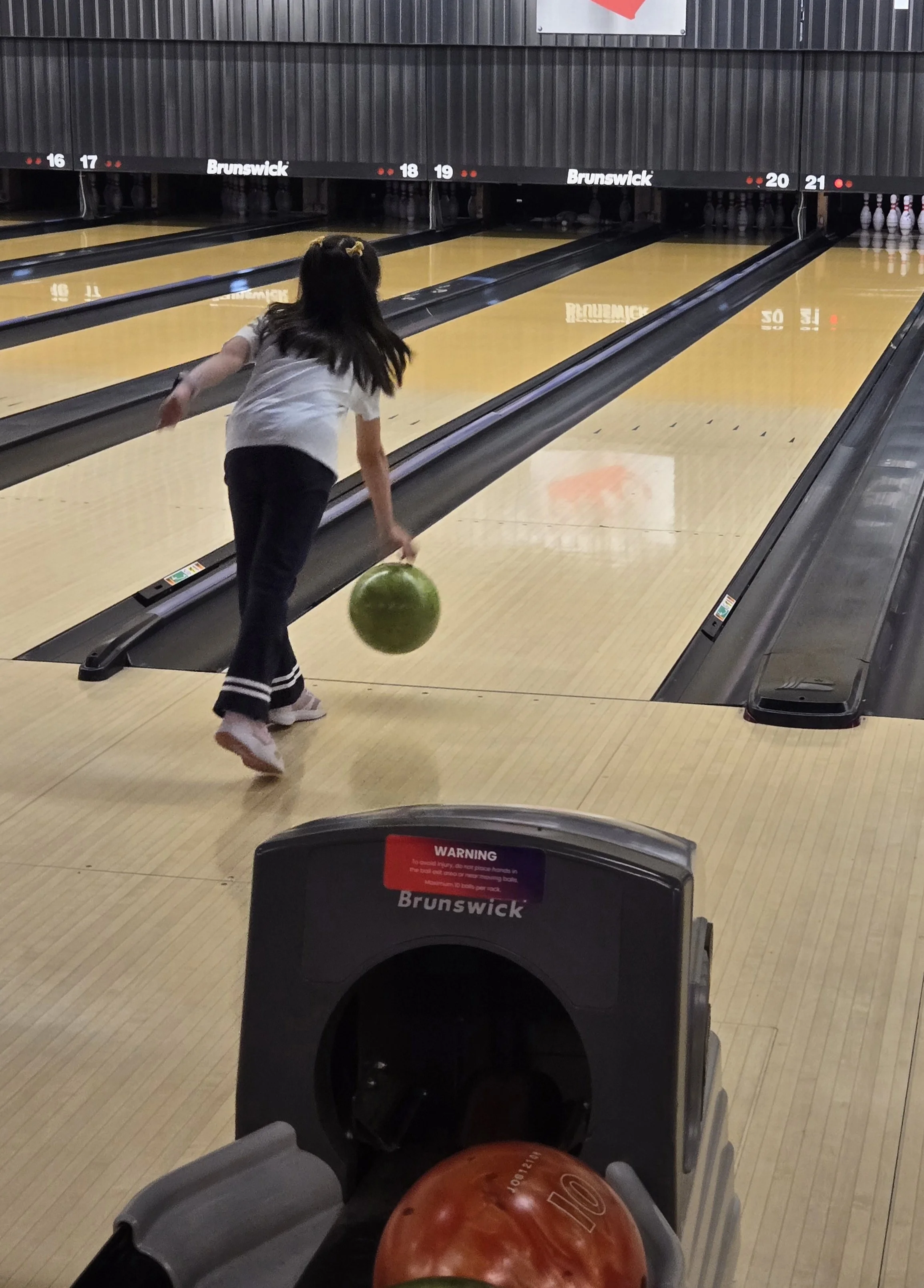 A young girl with long dark hair, wearing a white shirt and dark pants, is bowling at a bowling alley. She is about to release a green bowling ball down lane 18. The lanes are numbered 16 through 21, with pins visible at the end of lane 20.