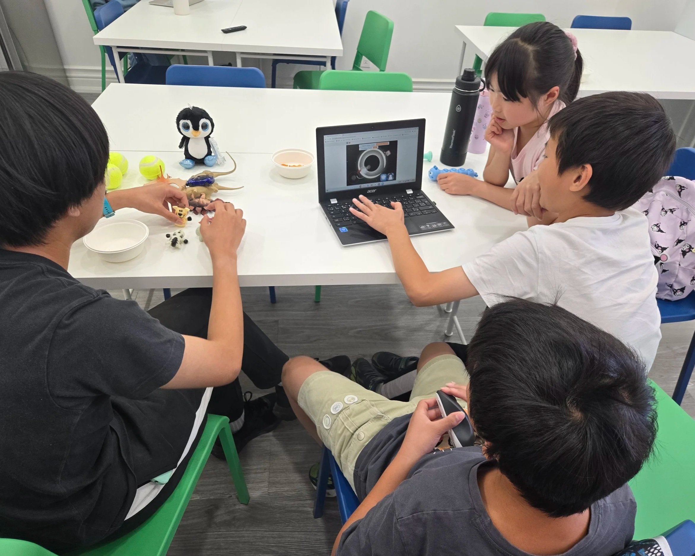 Group of children sitting around a white table watching a laptop screen, with toys and a stuffed penguin on the table.