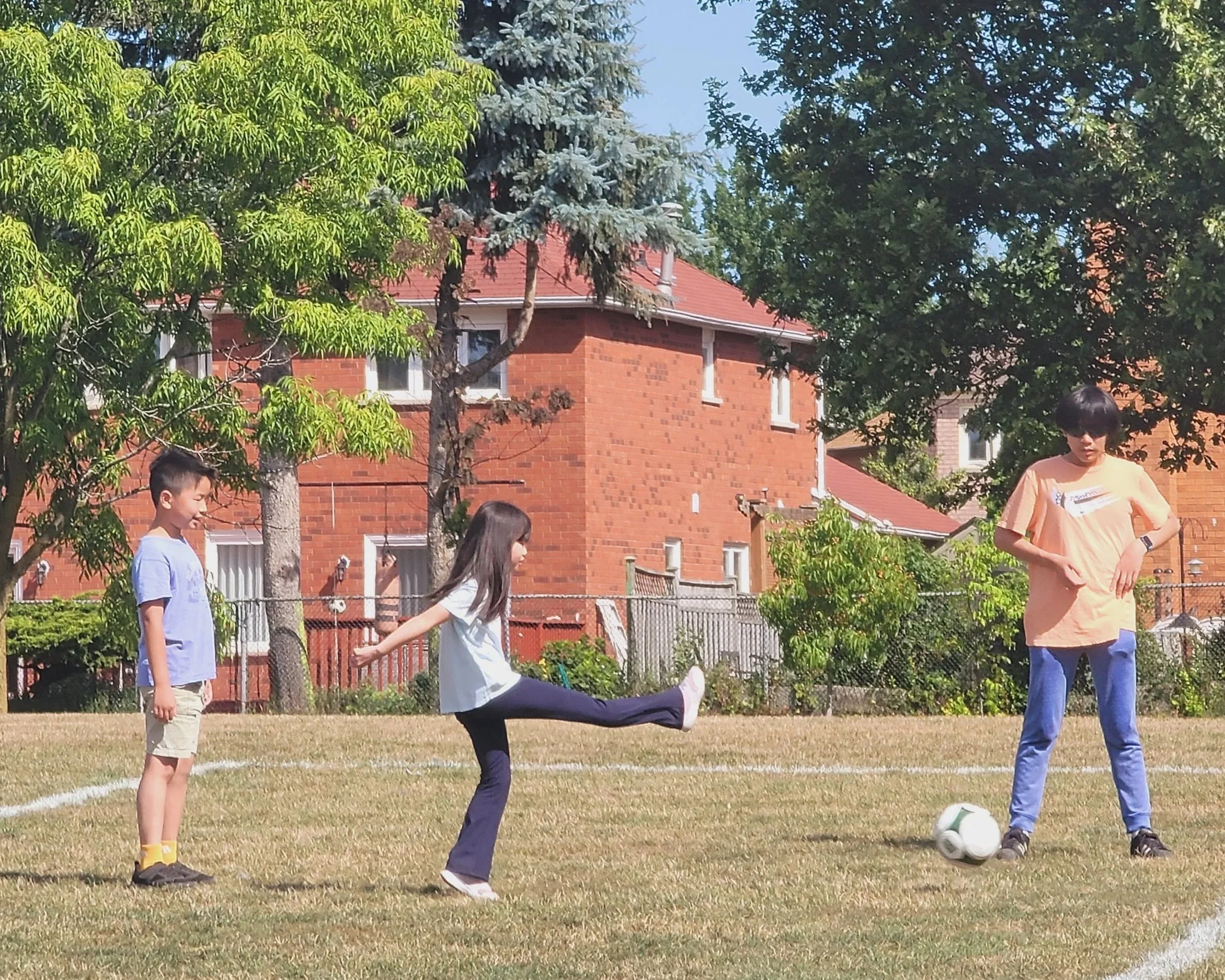 Three children playing soccer on a grassy field, with trees and red brick houses in the background on a sunny day.