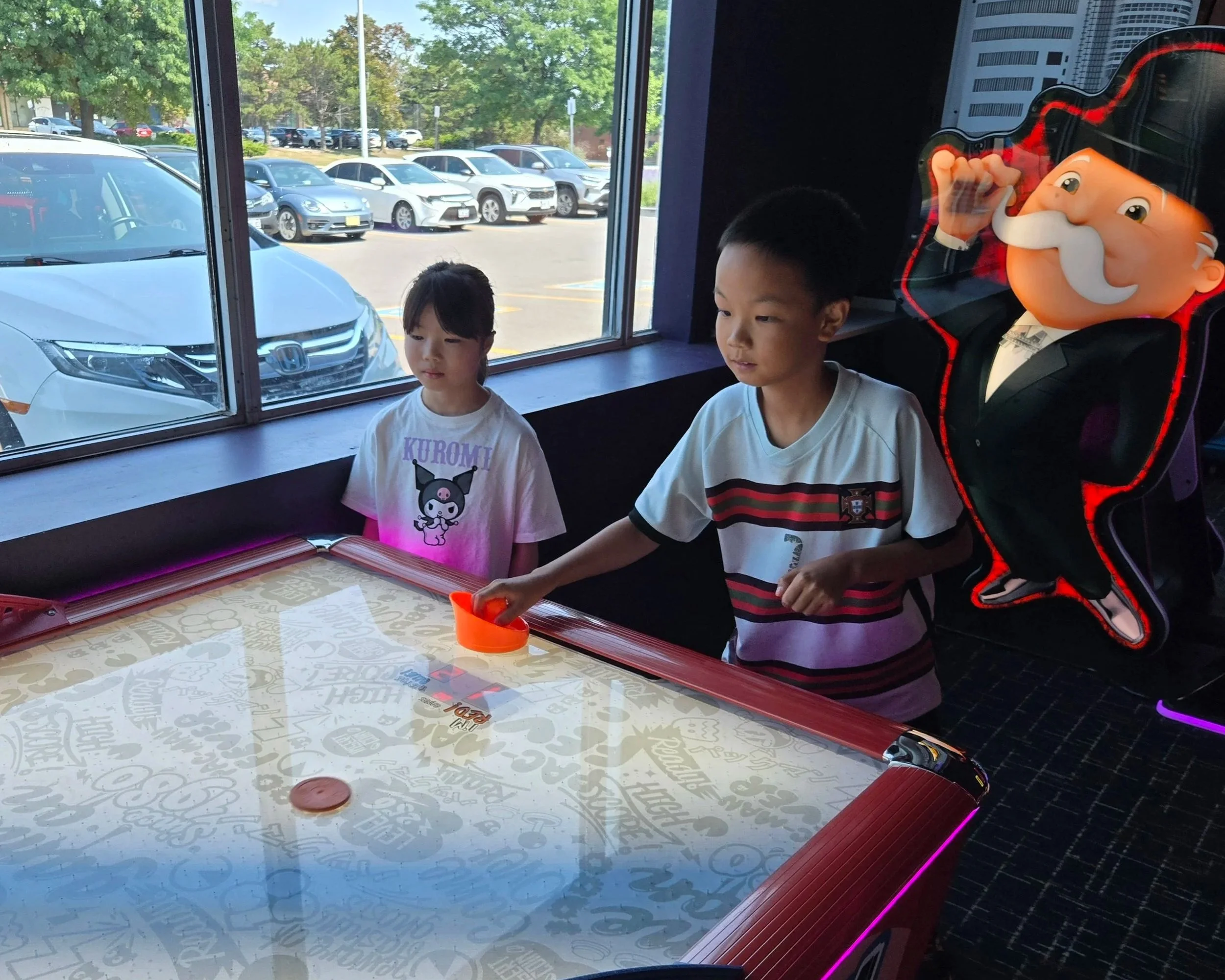 Two children playing air hockey at an indoor arcade, with a large Mario-themed balloon character in the background.