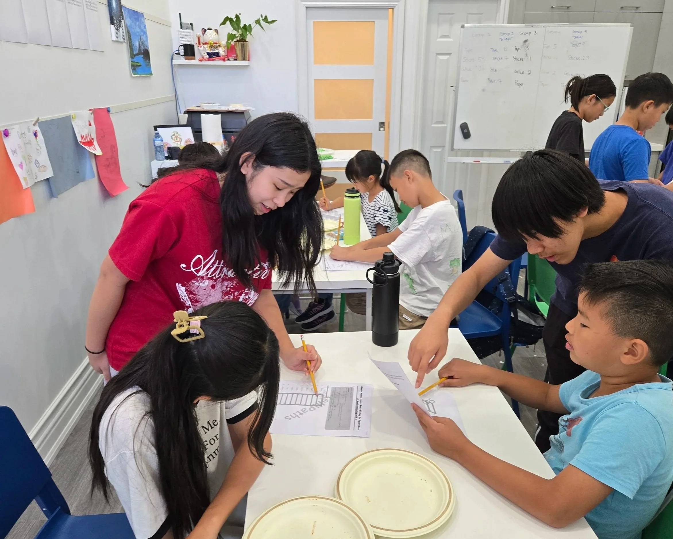 A classroom scene with children and adults engaging in an activity at a table. A woman in a red shirt is assisting a girl with long black hair and a hair clip. Other children are working on papers, some with pencils, while a man points at a person's paper. The walls of the room have artworks and a whiteboard in the background.