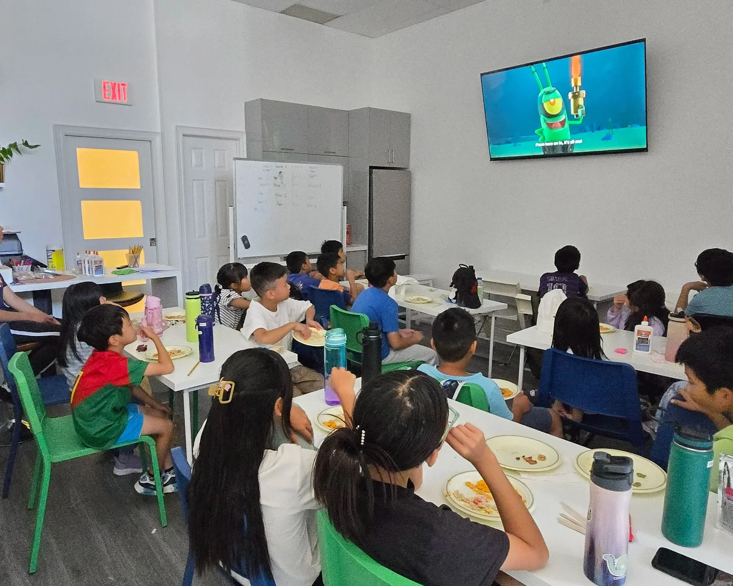 Children sitting at tables in a classroom watching an animated show on a large wall-mounted TV.