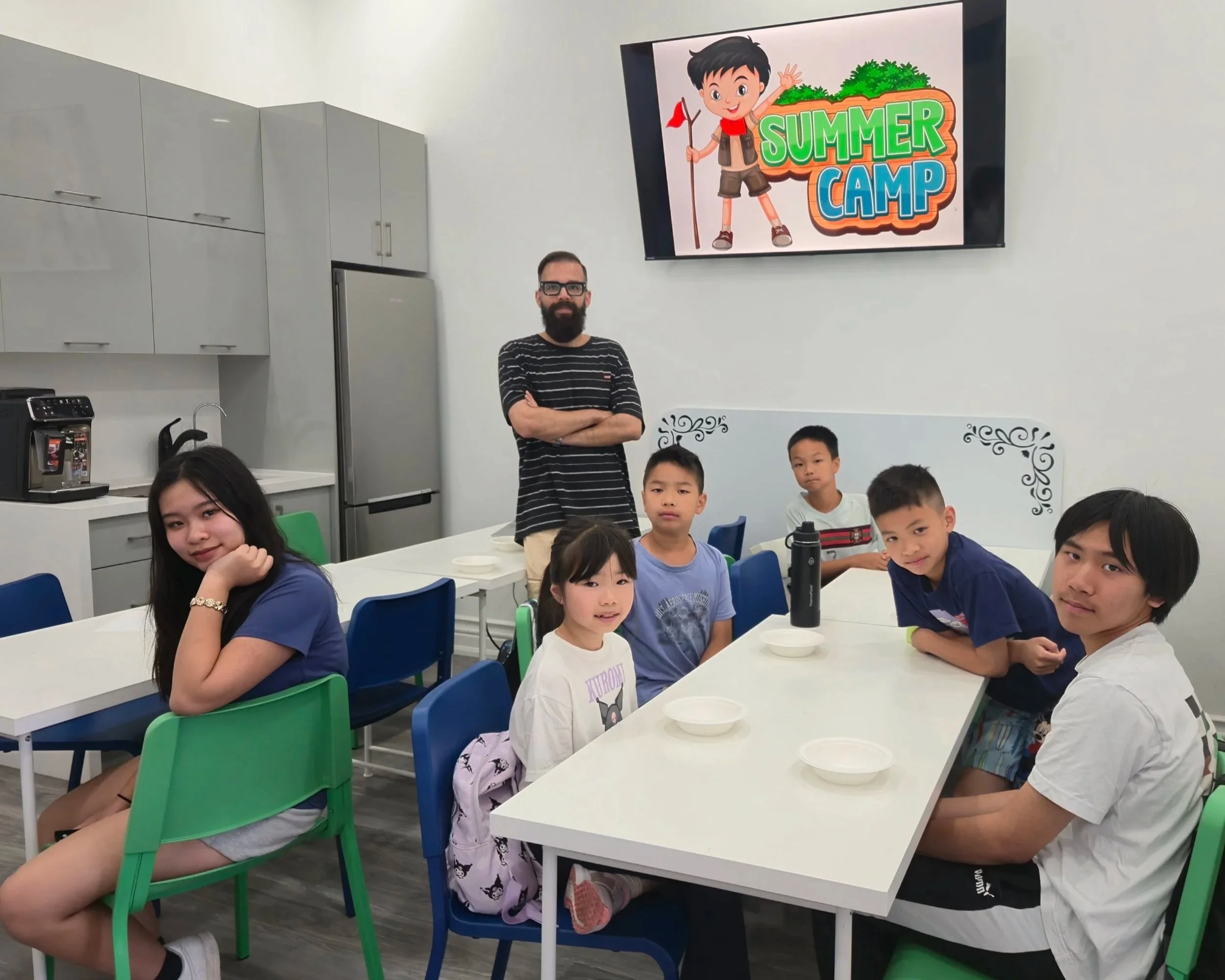 A group of children sitting at a table with some empty bowls, in a room with a kitchen background, with a man standing behind them, and a large screen on the wall displaying a 'Summer Camp' graphic.