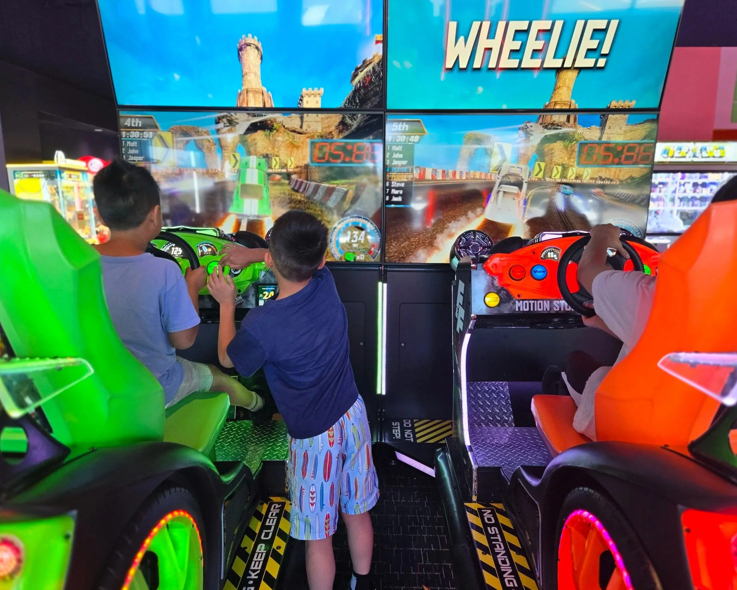 Two boys playing a racing arcade game at an arcade, with a third person steering on the right. The arcade machines have bright red and green seats, and large screens displaying a race car game with a castle and track visible.