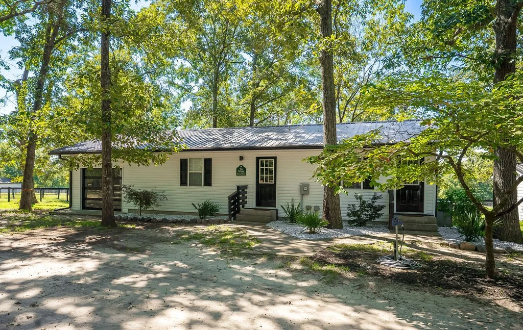 Single-story white house with black shutters, surrounded by trees and greenery, on a dirt yard with some shade.