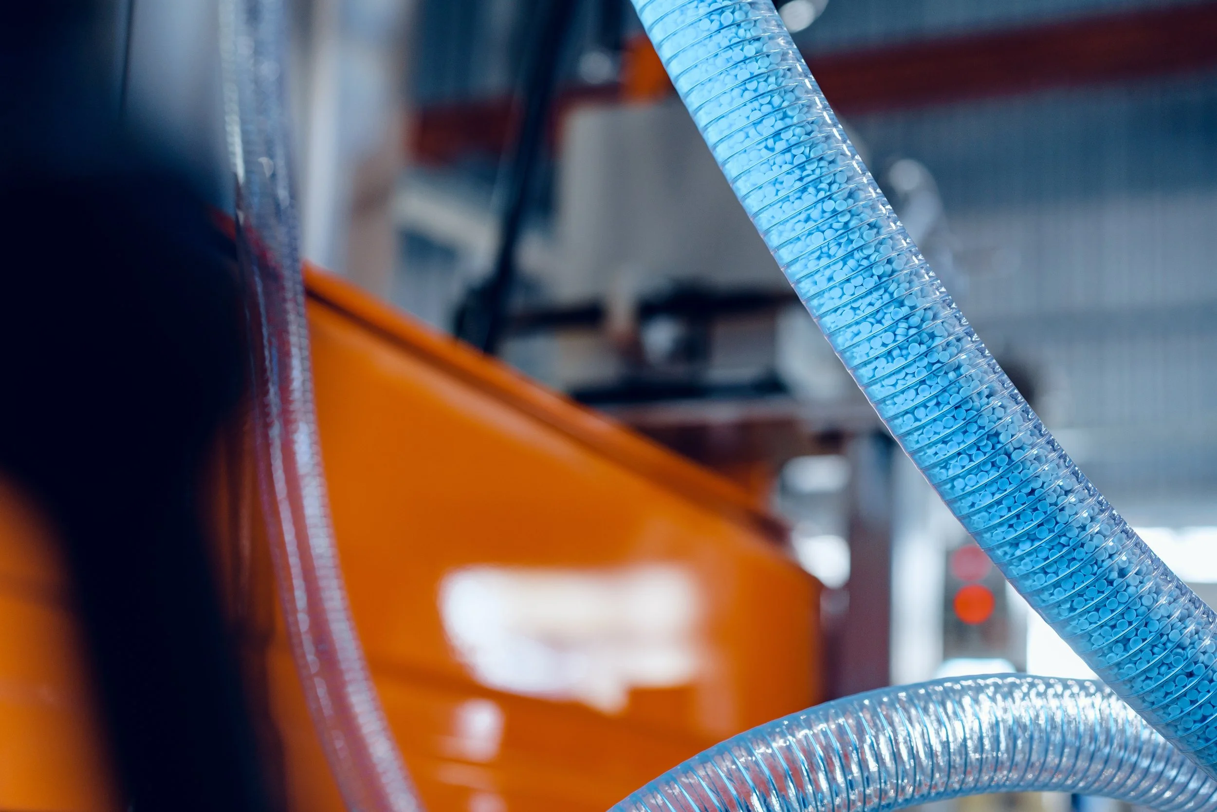 Close-up of blue corrugated hose connected to machinery in an industrial or workshop setting, with orange equipment and blurred background.