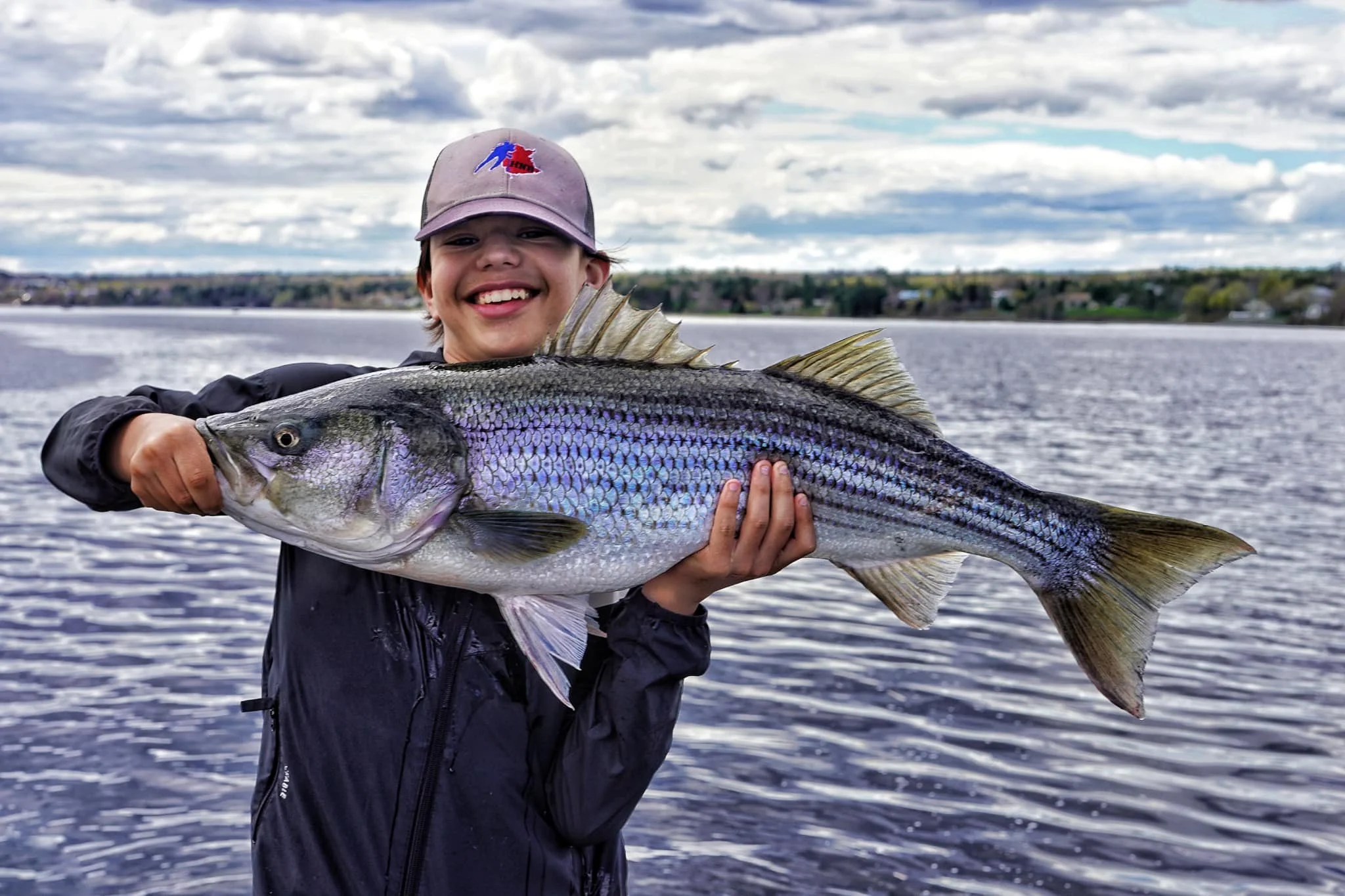 A young boy smiling and holding a large striped bass near the miramichi with a cloudy sky in the background.
