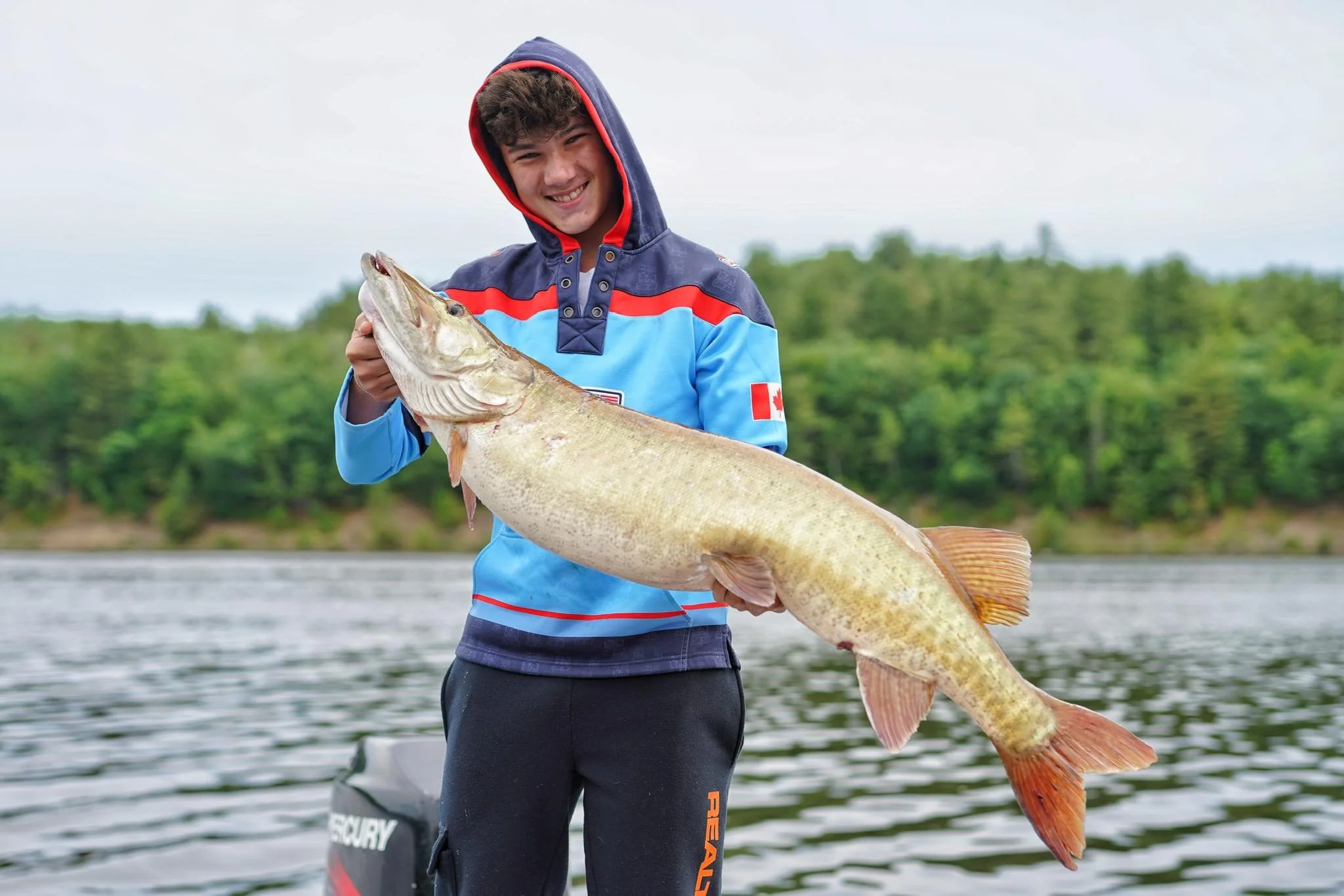 A young man wearing a blue and red hooded jacket holding a large muskie in a boat on a lake in woodstock New Brunswick, with a background of trees and overcast sky.