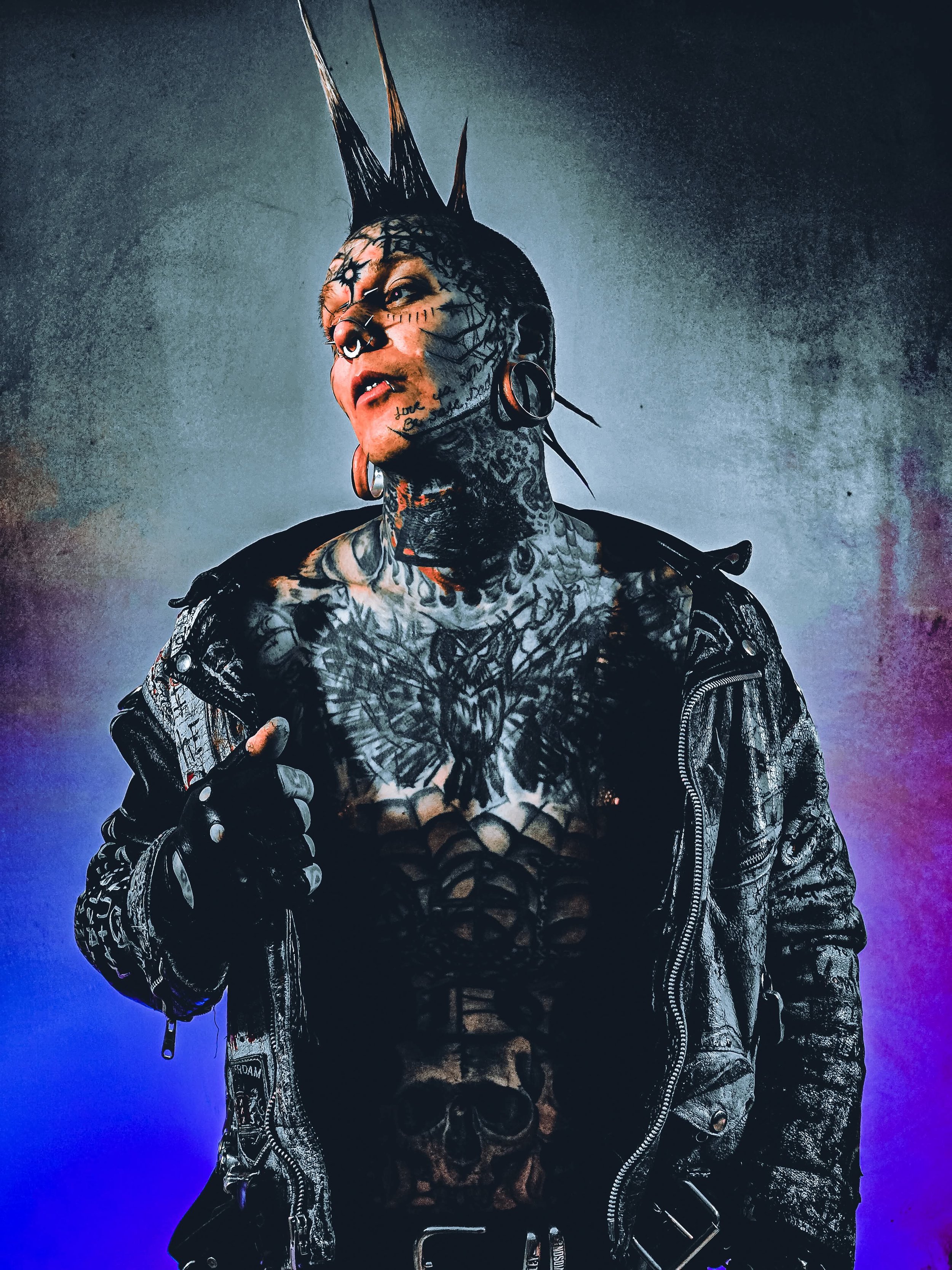 A person with multiple tattoos, a mohawk hairstyle, piercings, and a leather jacket, posing against a colorful gradient background.