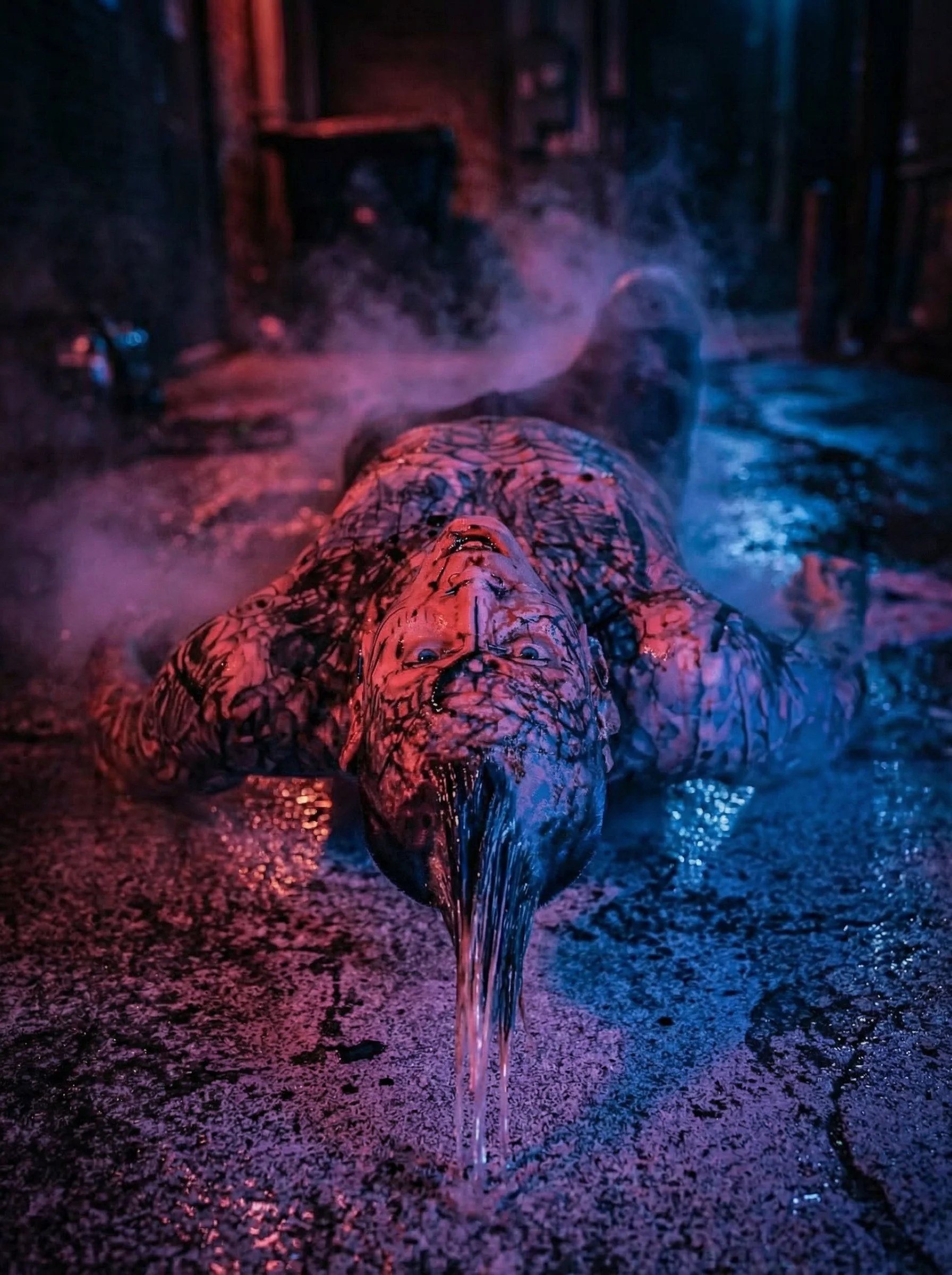 A person lying on the ground with their face and body covered in black paint or ink, emitting smoke or fog, in a dimly lit room with colored lights creating a moody, surreal atmosphere.