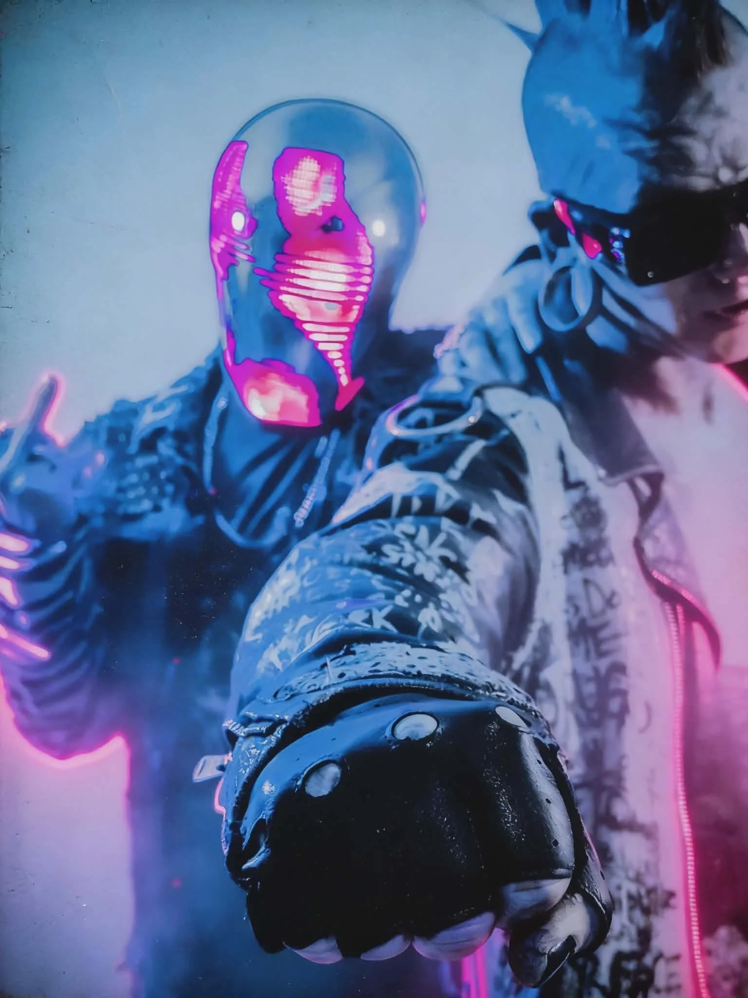 Person wearing sunglasses and a hooded jacket, with a glowing pink and blue mask or helmet, reaching out towards the camera.