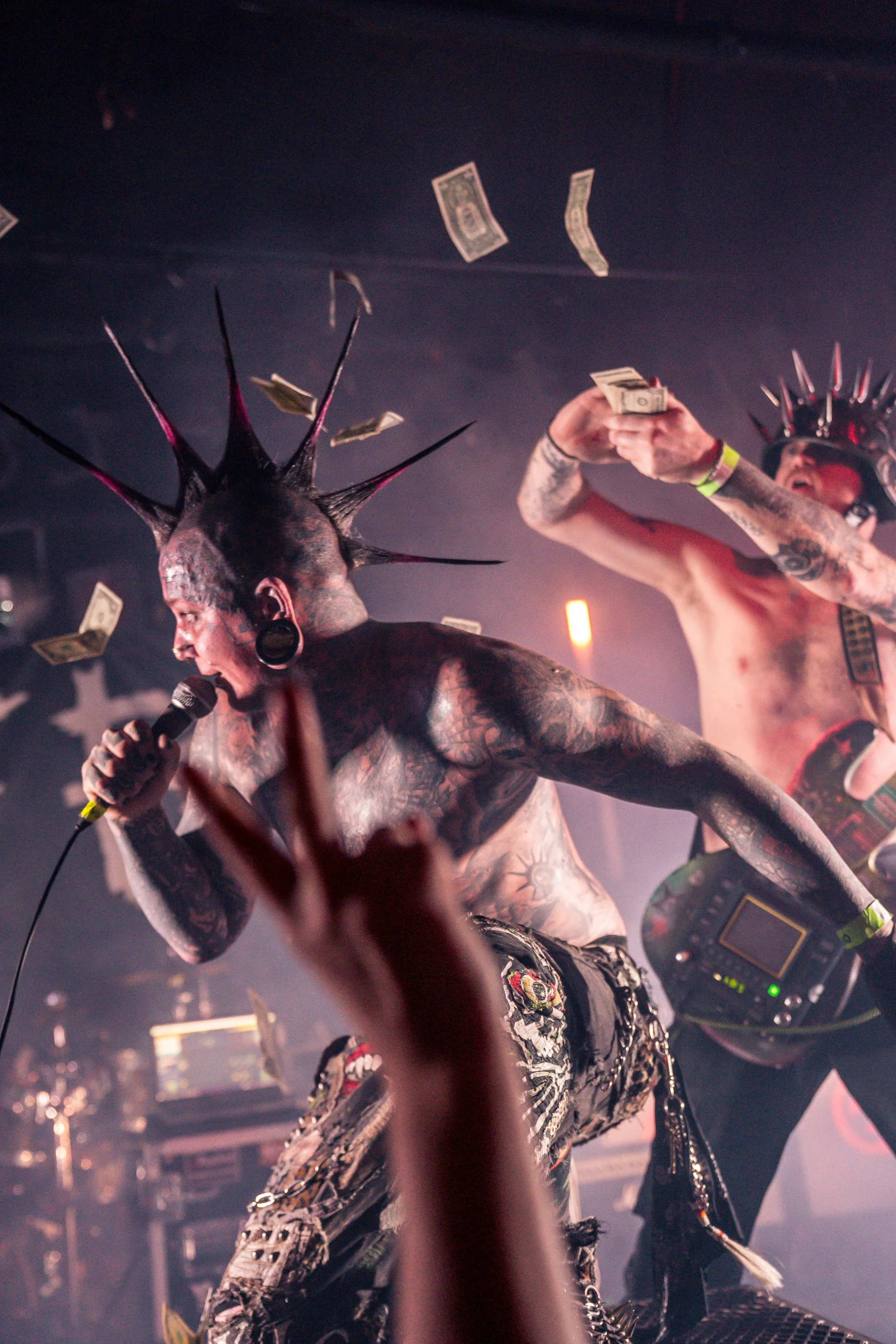 Tattooed performers with piercings and spiked headwear on stage, one singing into a microphone and the other playing a guitar, with dollar bills falling from above in a concert setting.
