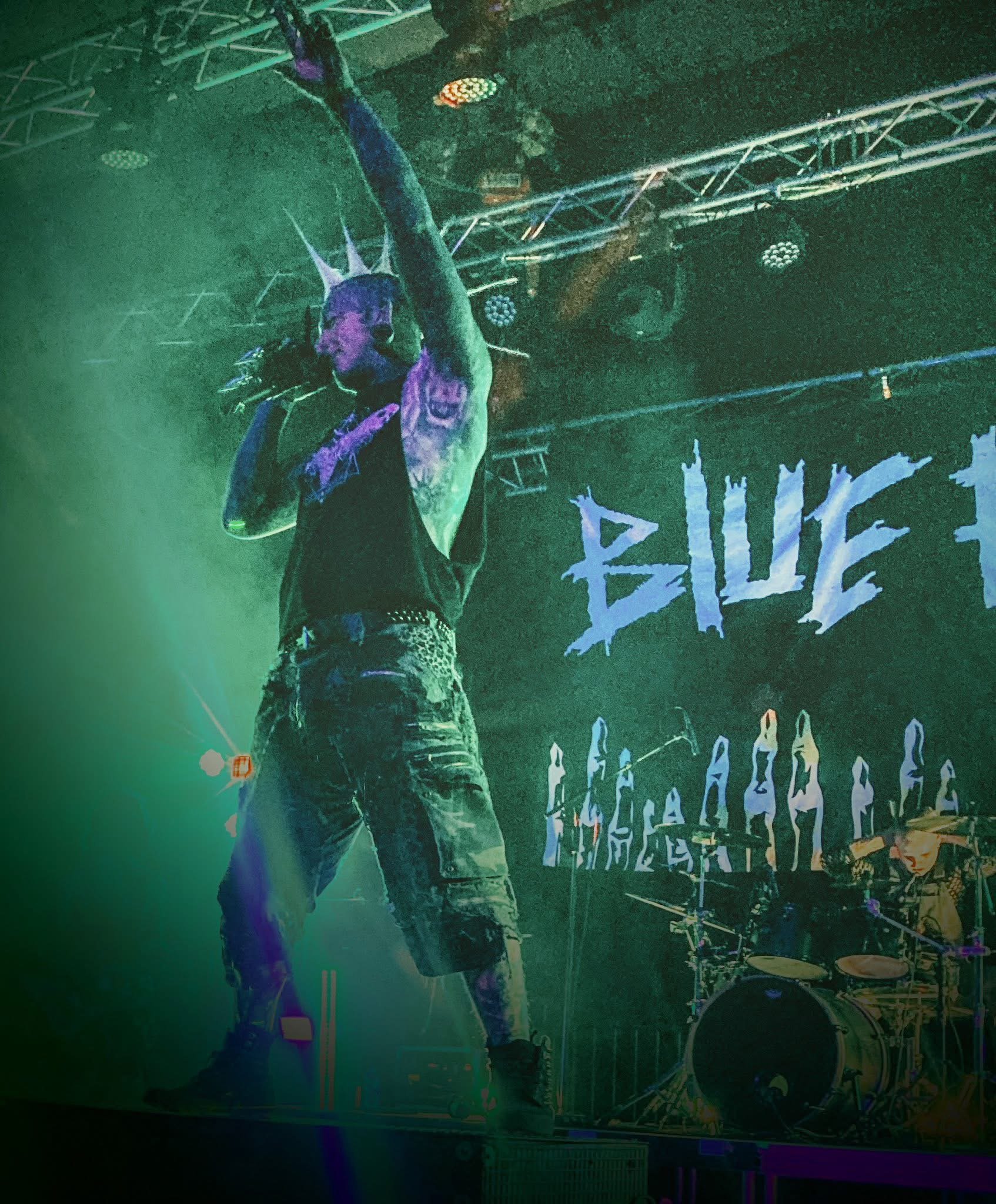A performer with face paint, wearing a crown on their head, holding a microphone, on a stage with green lighting. A drum set is behind them, and a backdrop displays the word "BLUE" in stylized text.