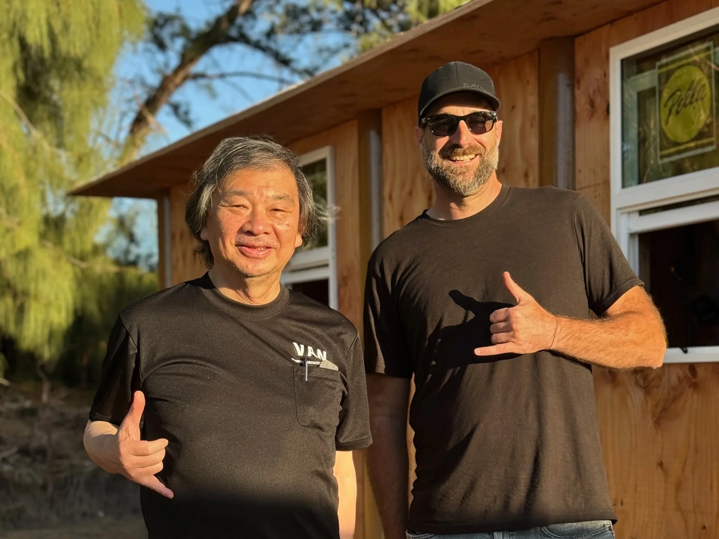 Two men standing outside in front of a wooden building, smiling and making hand gestures. One man is Asian with gray hair, wearing a black T-shirt. The other man is Caucasian with a beard, wearing sunglasses, a black baseball cap, and a black T-shirt.