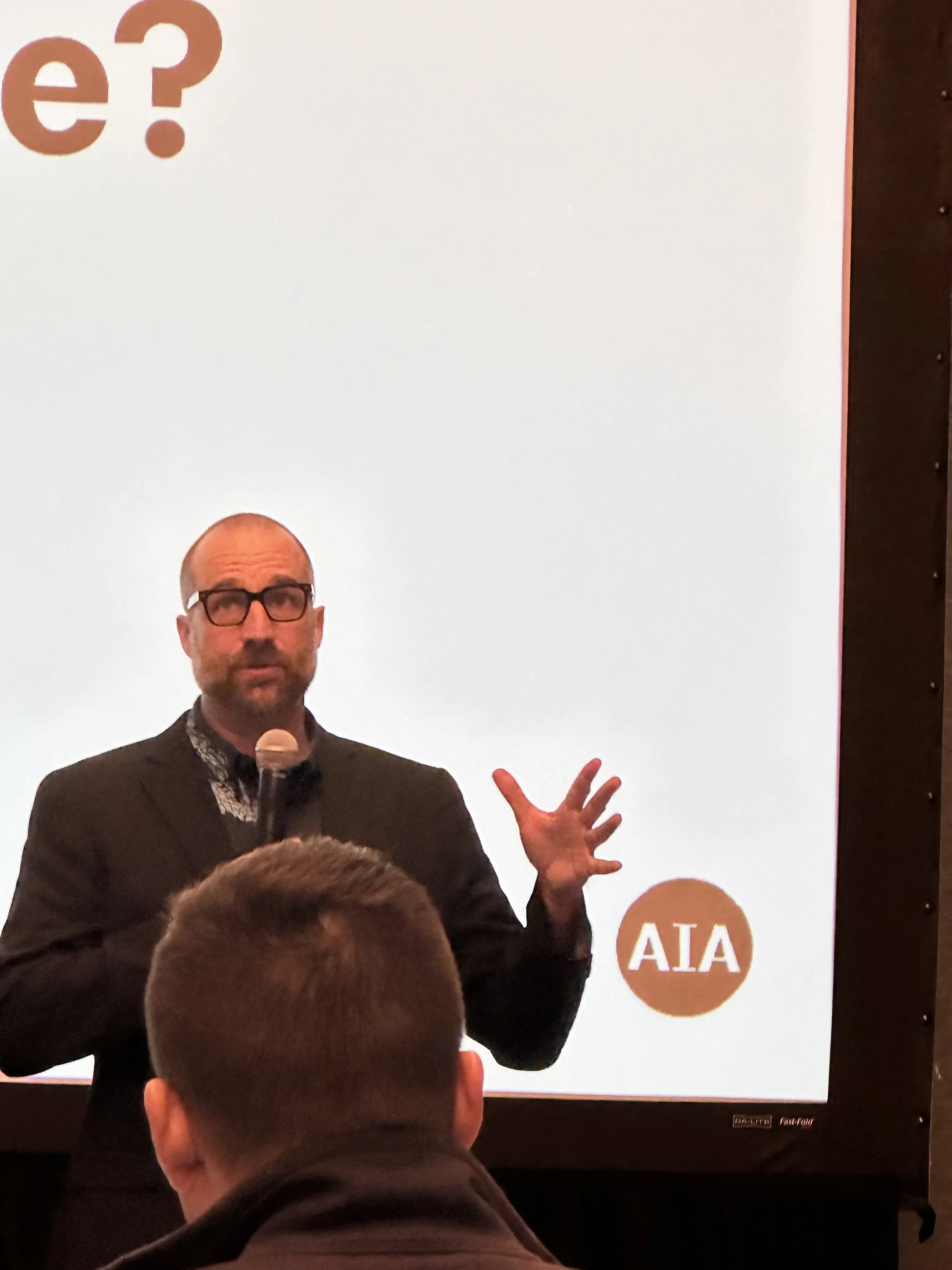 A man wearing glasses, dark blazer, and patterned shirt holding a microphone, giving a presentation in front of a white screen with the letters 'e?' in the upper left corner and the logo 'AIA' in the lower right corner. An audience member with short brown hair is in the foreground.