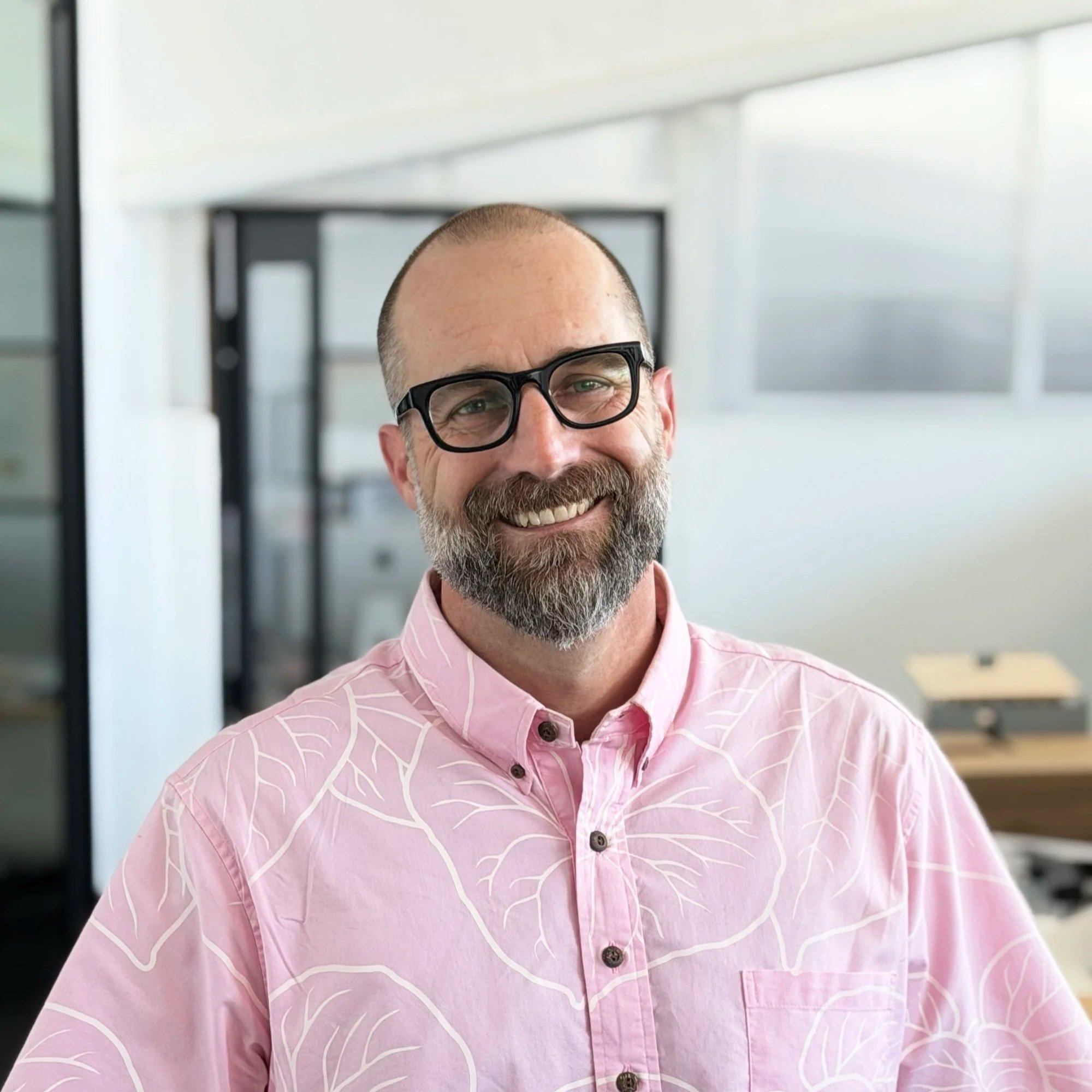 A smiling man with glasses and a beard, wearing a pink shirt with a white leaf pattern, in an office setting with large windows and modern decor.