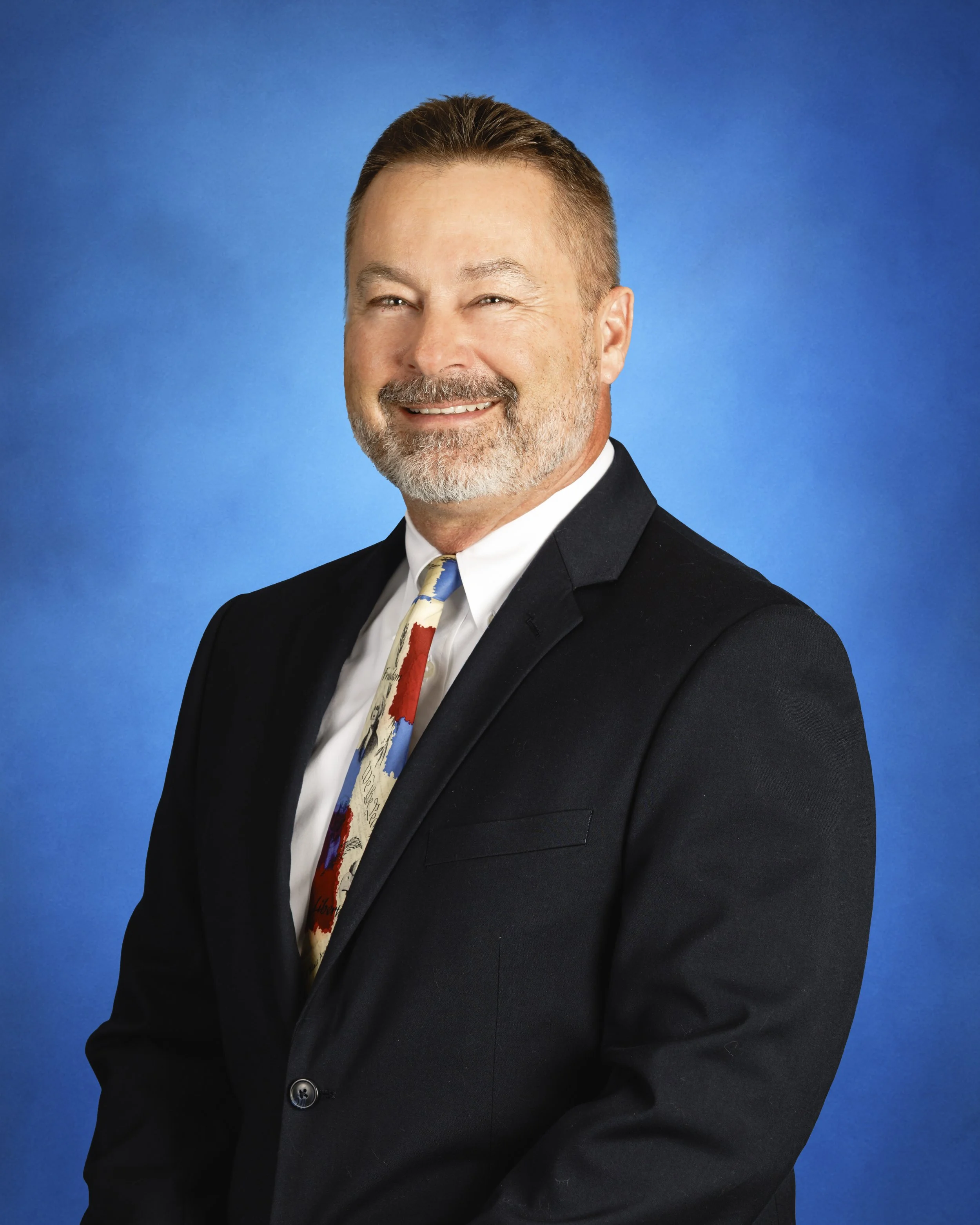 A middle-aged man with a beard and mustache, wearing a black blazer, white shirt, and a colorful tie, smiling against a blue background.