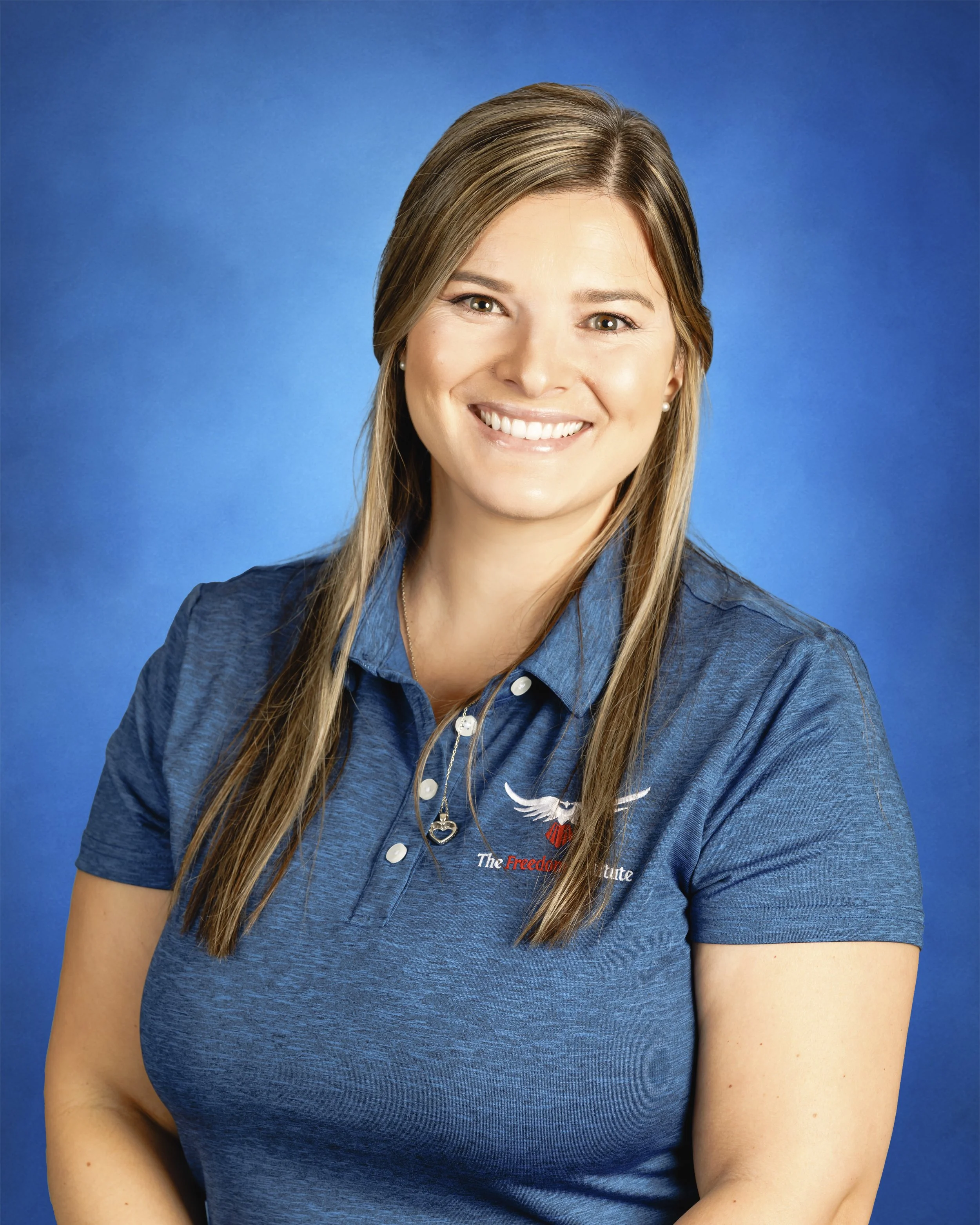 A woman with long brown hair smiling, wearing a blue polo shirt with a logo and a necklace, against a blue background.