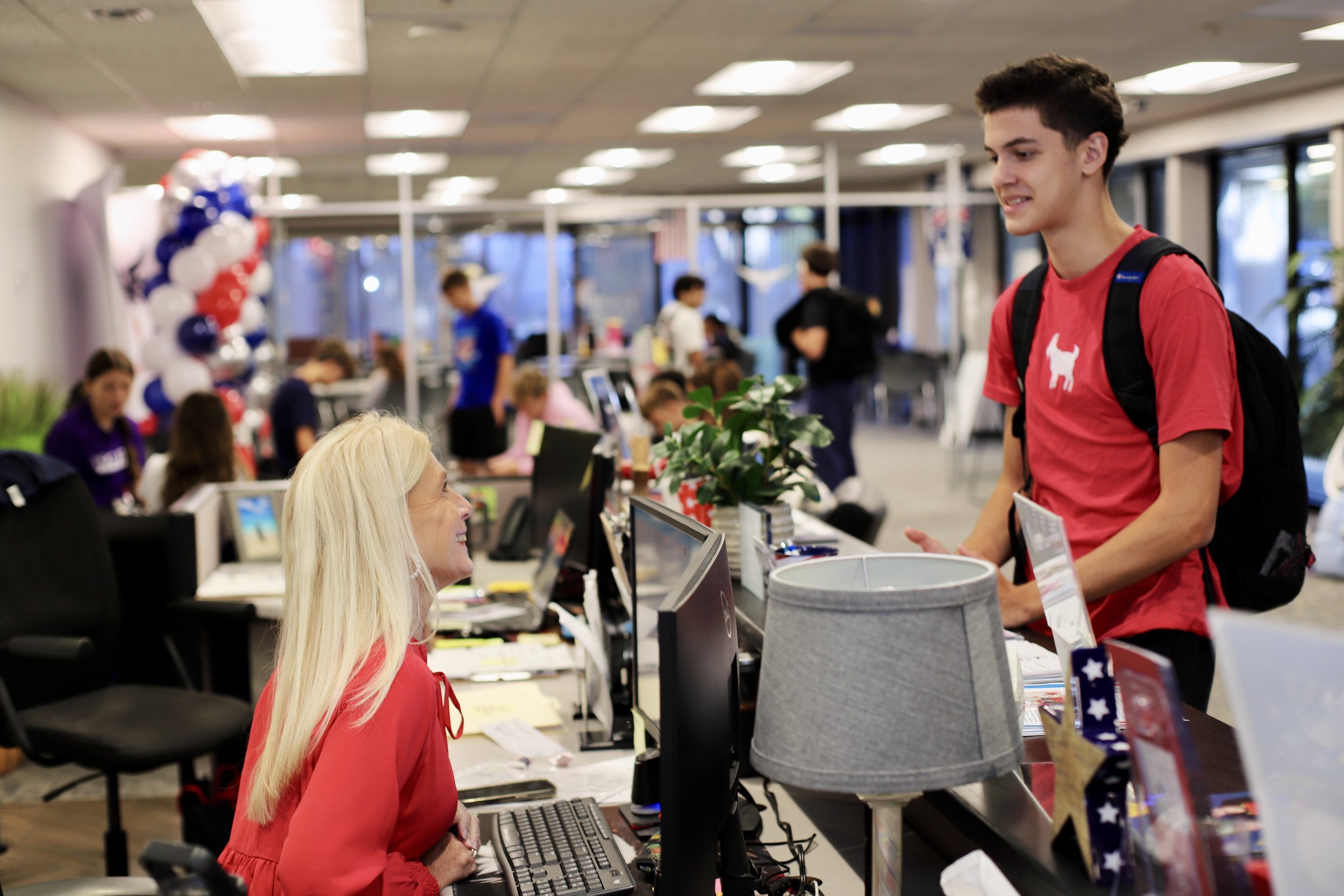 A woman with long blond hair wearing a red blouse is smiling and talking to a young man with dark hair and a red shirt with a white dog silhouette, at a reception desk inside an office or community center decorated with red, white, and blue balloons.