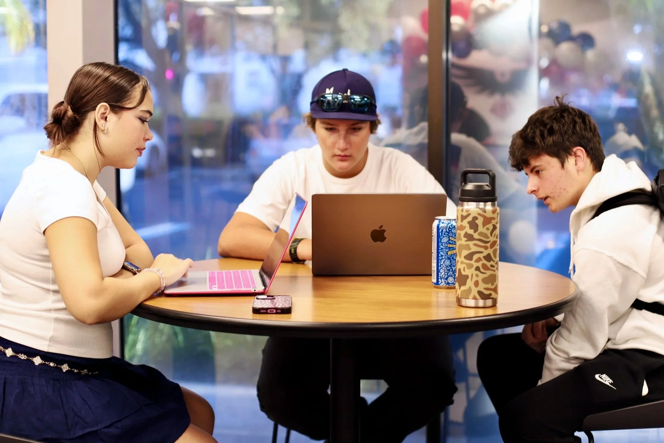 Three young people sitting around a round wooden table at a cafe, each using a laptop or smartphone, with drinks on the table and large windows in the background.