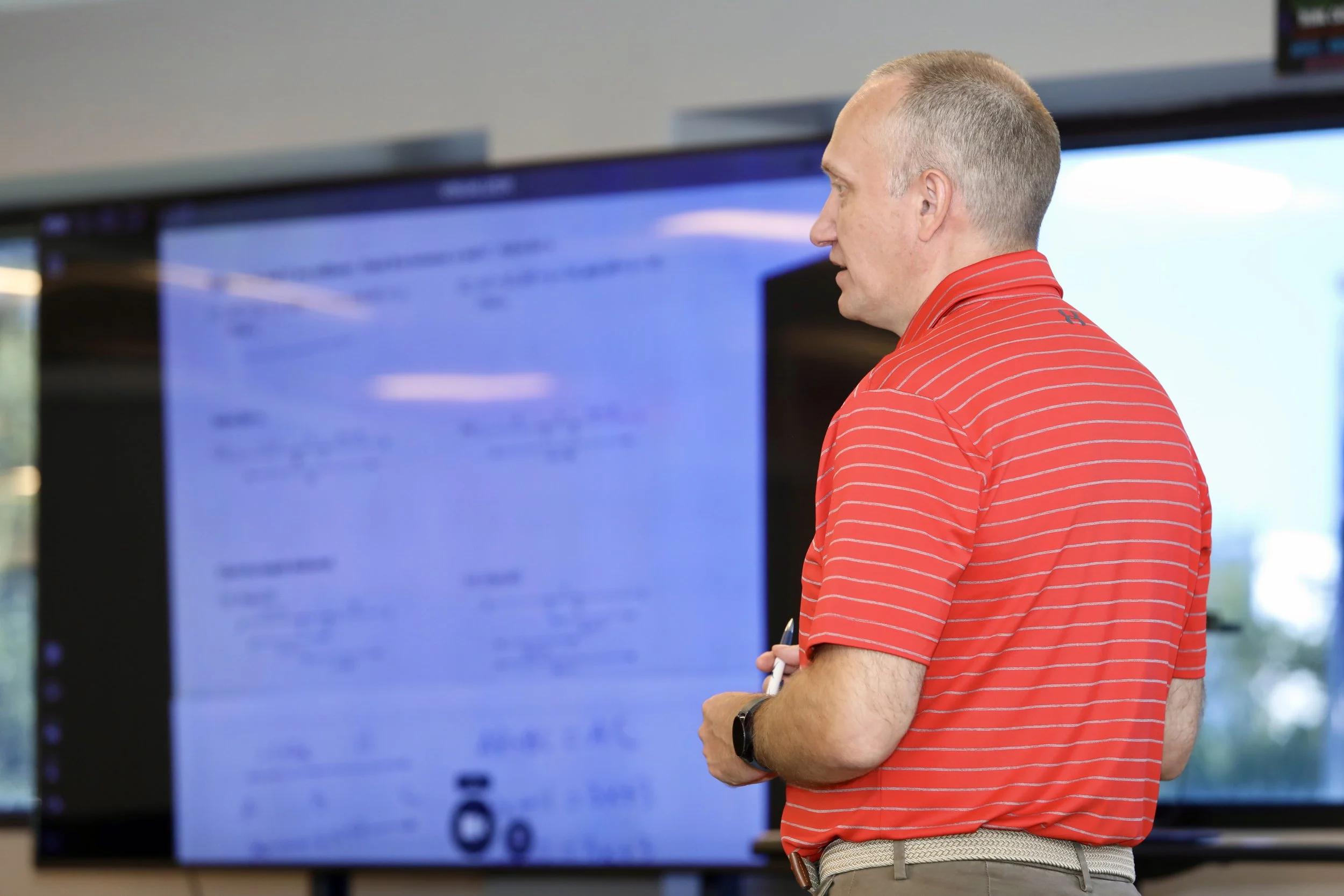 A man with a shaved head in a red striped polo shirt holding a pen, standing in front of a large screen displaying text during a presentation or lecture.