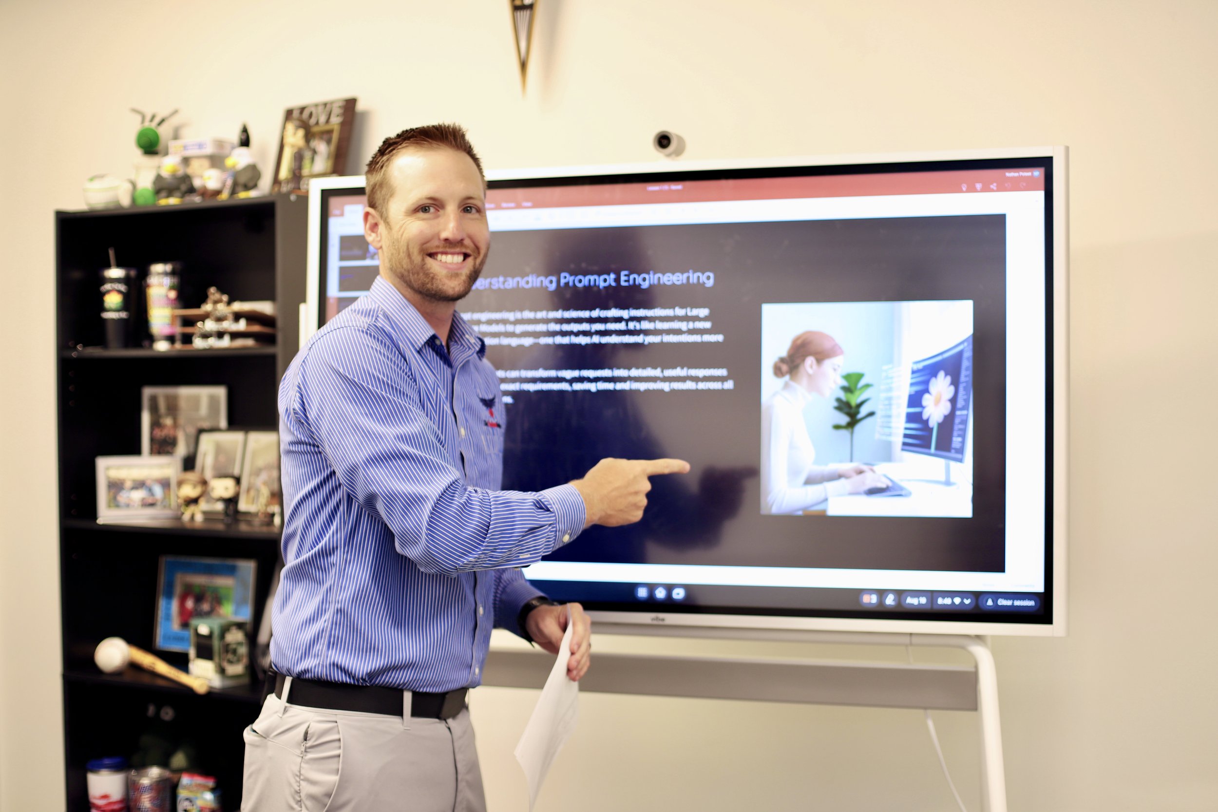 Man in striped shirt pointing at presentation slide on digital screen in a room with black bookshelf filled with framed photos and items.