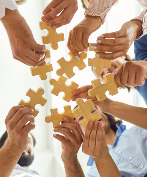 9 hands each holding a wooden puzzle piece trying to come together