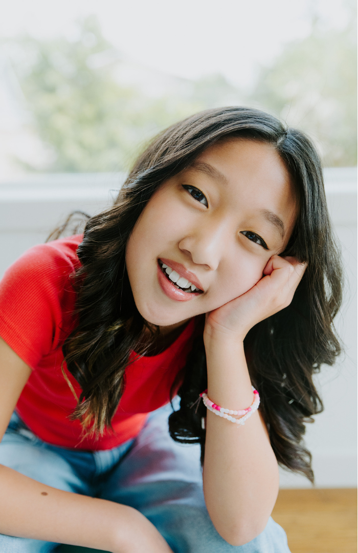 Smiling girl with long, wavy dark hair wearing a red shirt and hair bracelets, resting head on hand, indoors with a bright window behind.