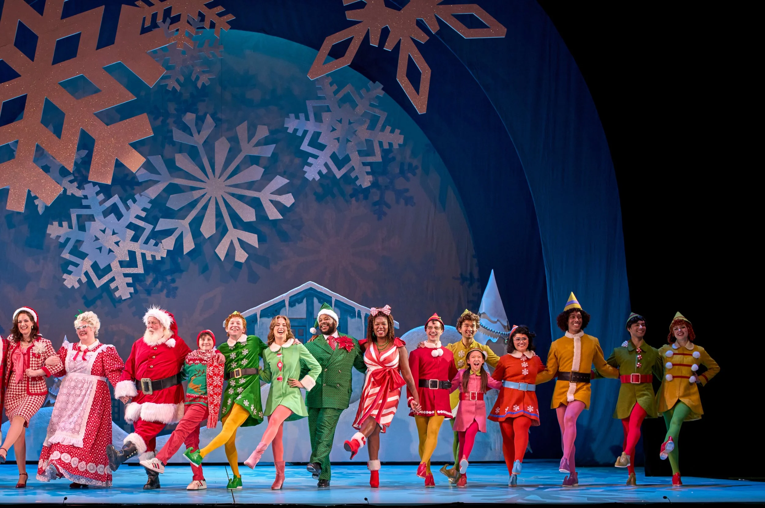 People dressed in colorful holiday costumes performing a choreographed dance on stage with large snowflake decorations and a winter cityscape backdrop.