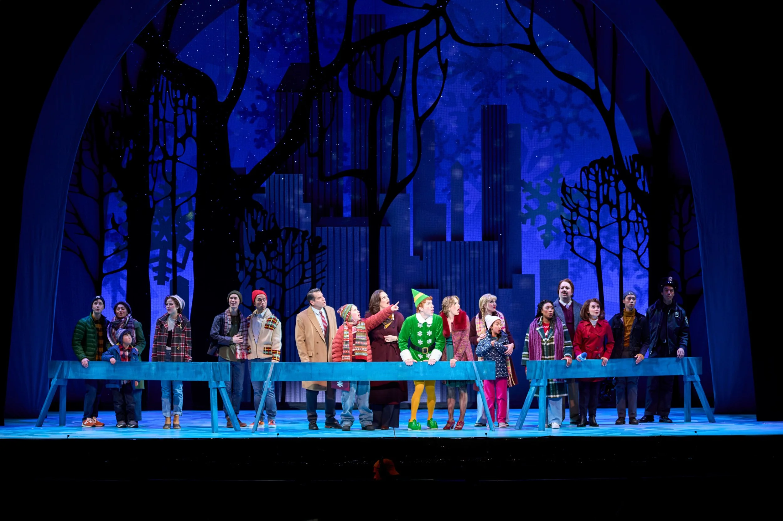 A group of people on stage performing in a holiday play, dressed in festive and winter clothing, with a winter cityscape backdrop and decorated trees.