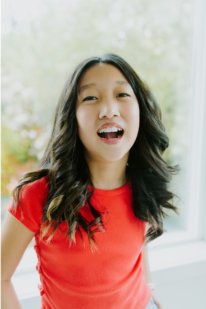 A young girl with long, wavy dark hair wearing a red t-shirt, smiling and looking directly at the camera with her mouth open. She is standing in front of a window with blurred greenery outside.