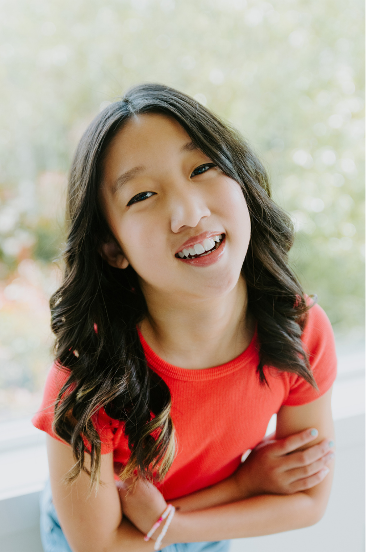 A young girl with long black wavy hair, wearing a red shirt, smiling playfully with her arms crossed.