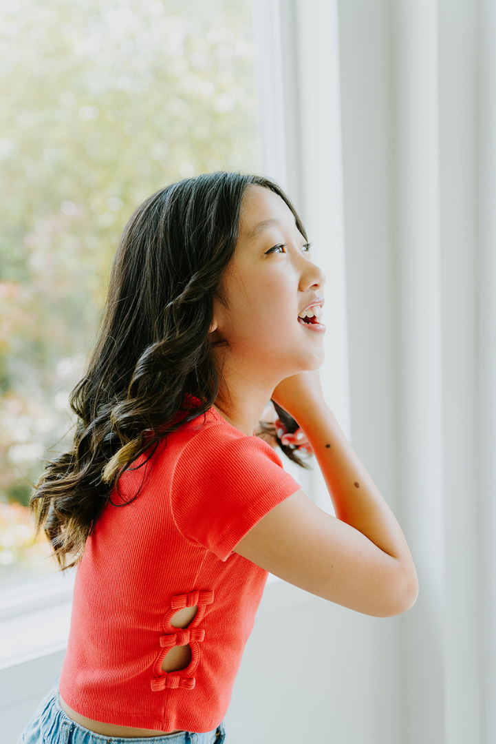 A girl with wavy dark hair looking out of a window, smiling, wearing a red top and light-colored bottoms.