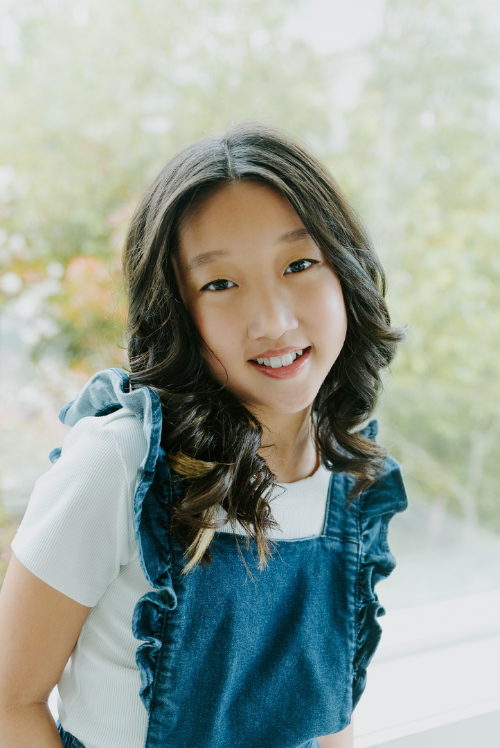 A young girl with long, wavy dark hair and a smile, wearing a white shirt and blue denim dress with ruffled shoulder straps, standing in front of a window with blurred green trees outside.