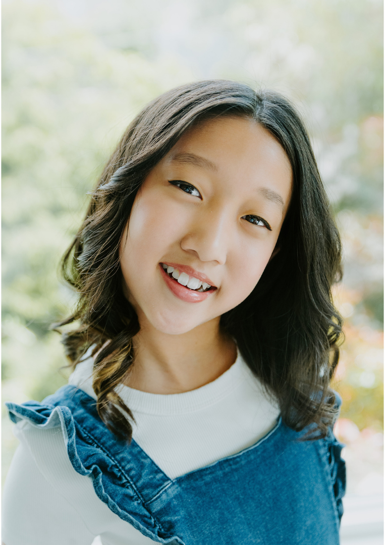 Close-up of a young Asian woman smiling outdoors with blurred green foliage in the background.