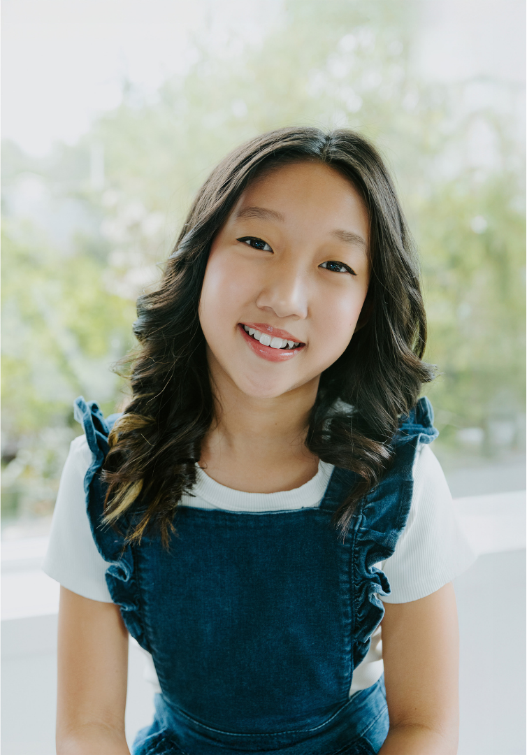 Young girl with long wavy dark hair smiling indoors with a window and green trees in the background.