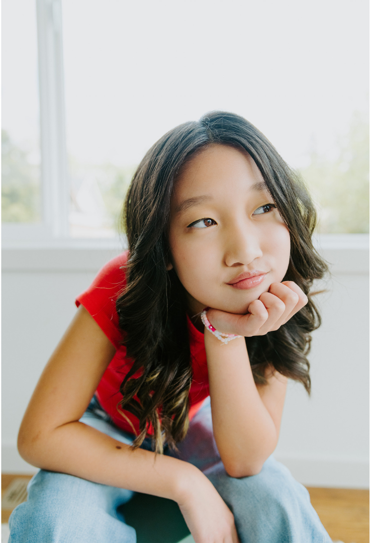 Young girl with long, wavy dark hair wearing a red shirt and blue pants, resting her chin on her hand while sitting indoors near a window.