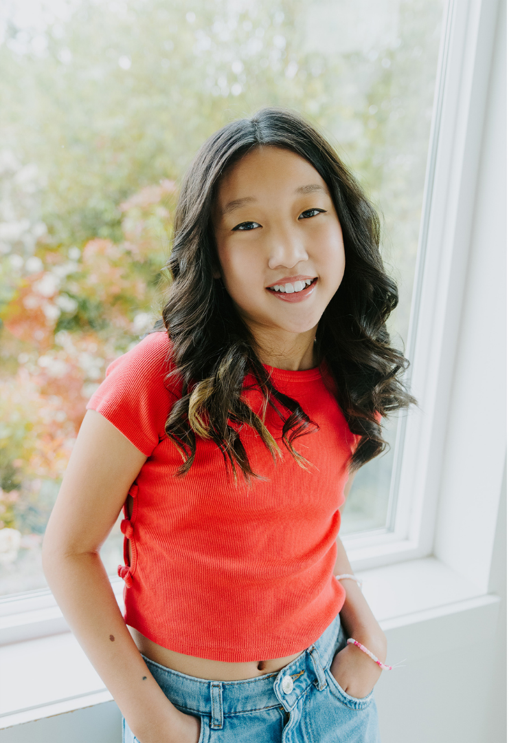 A young girl with long, wavy black hair smiling at the camera while standing indoors near a window with an outdoor view of colorful fall trees.