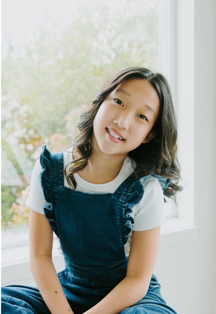 A young girl with shoulder-length wavy dark hair, smiling and looking at the camera, sitting near a window with a blurred view of trees outside.