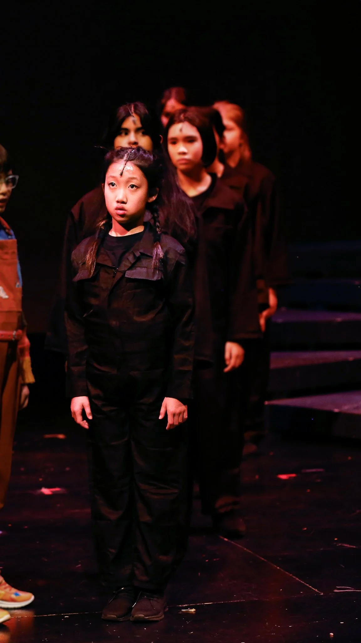 A group of children standing in a line on a stage, dressed in black clothing, with serious expressions.