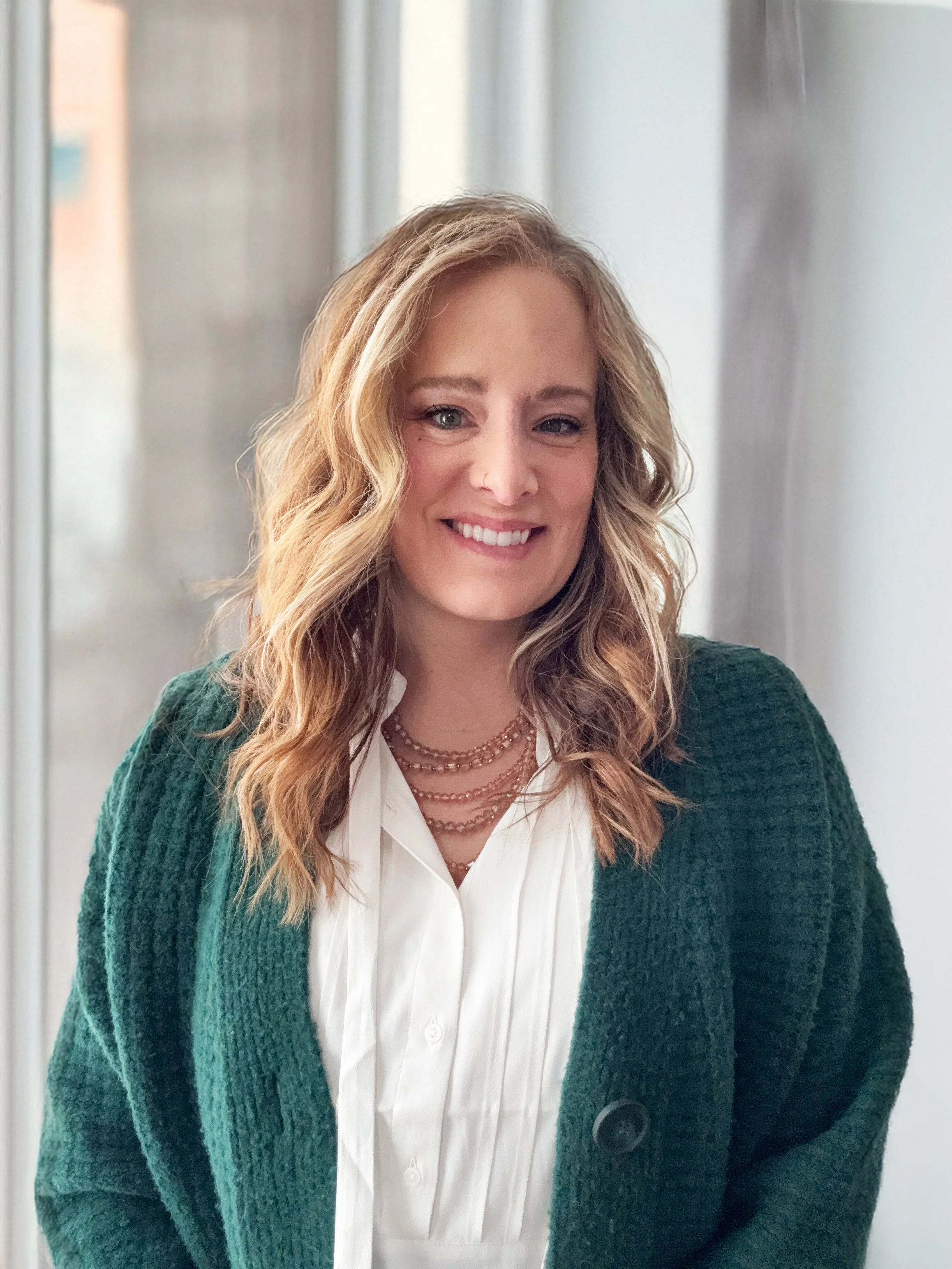 A smiling woman with wavy blonde hair wearing a white shirt, layered necklaces, and a green cardigan standing indoors near a window.