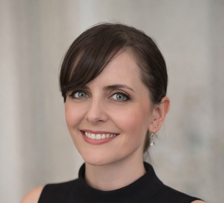 A smiling woman with short brown hair, blue eyes, and fair skin, wearing a black sleeveless top and earrings, standing in front of a neutral background.