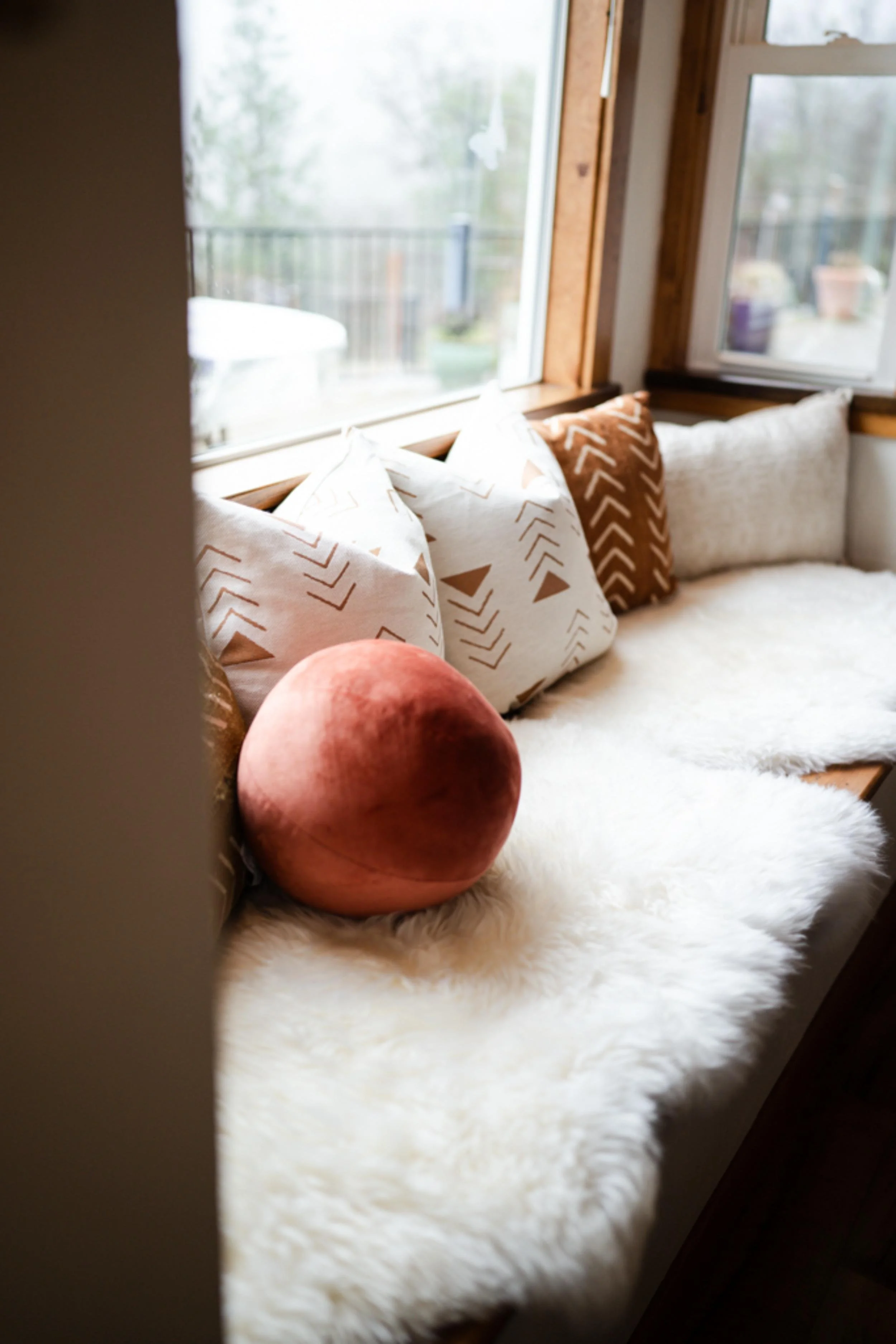 Sunlit window seat with white and brown patterned pillows, a round pink velvet cushion, and a white fluffy blanket.