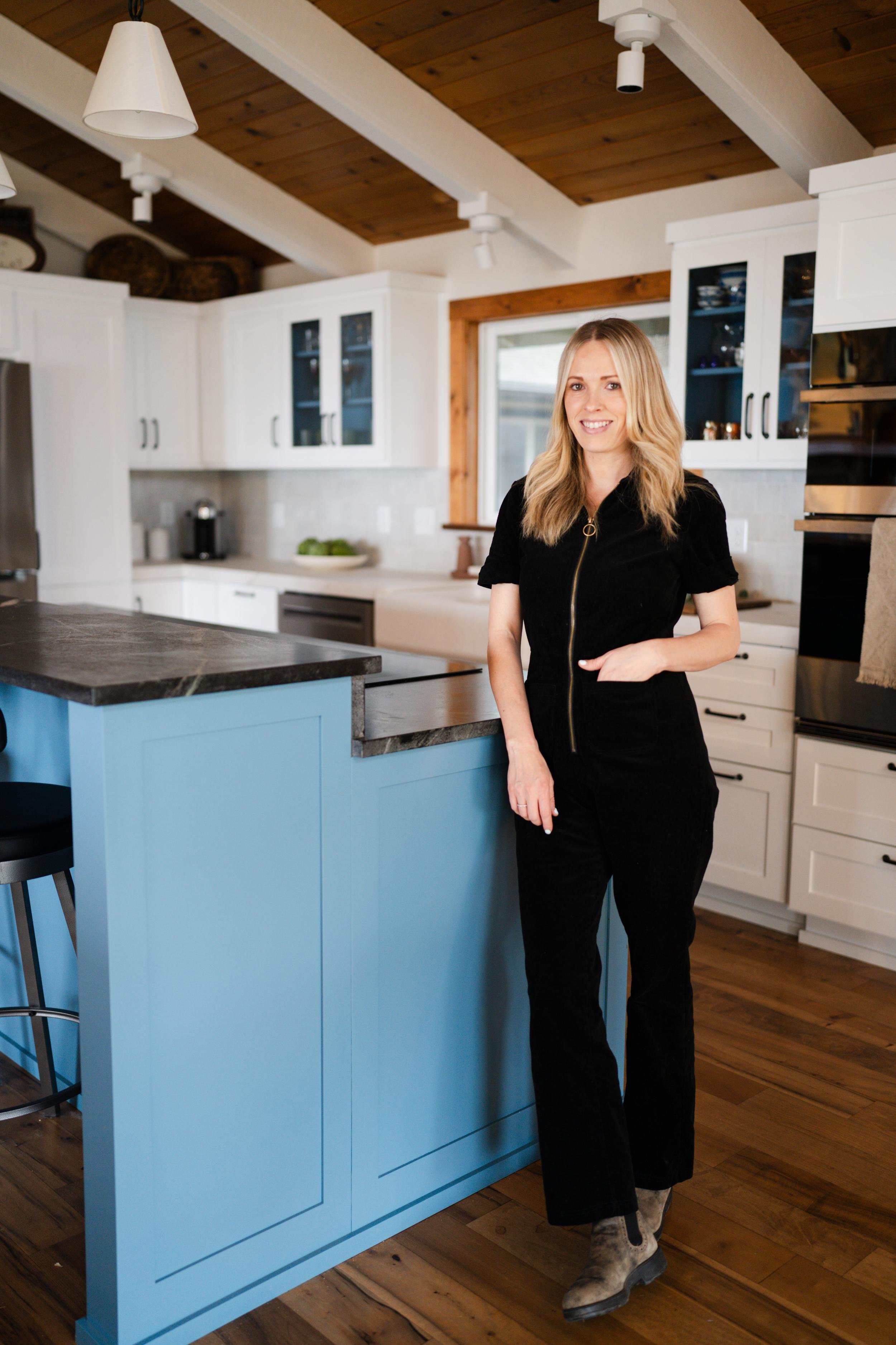 A woman in black clothing standing in a modern kitchen with white cabinets and a blue island, smiling.