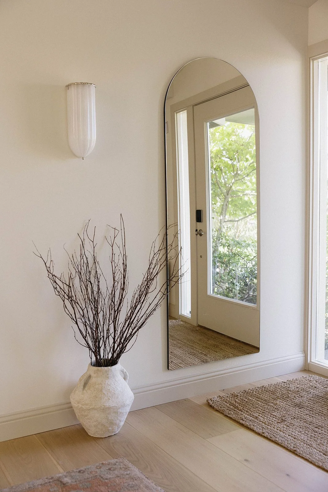 A hallway with a large, oval mirror on the wall, a white textured vase with branches on the floor, a wall sconce, and a glass door leading to a view of trees outside.