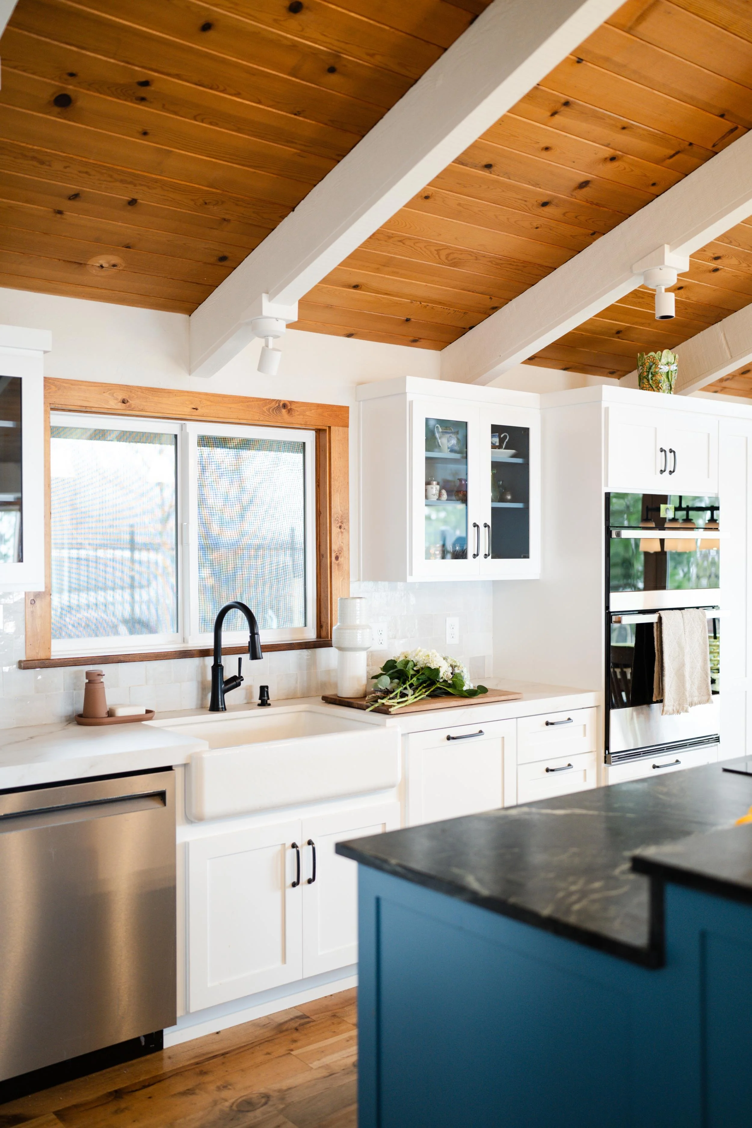 A modern kitchen with white cabinets, a farmhouse sink, and a window with wooden trim. The ceiling is made of wooden planks, and nearby there is a blue kitchen island with a black countertop. The space is decorated with some flowers and kitchenware.