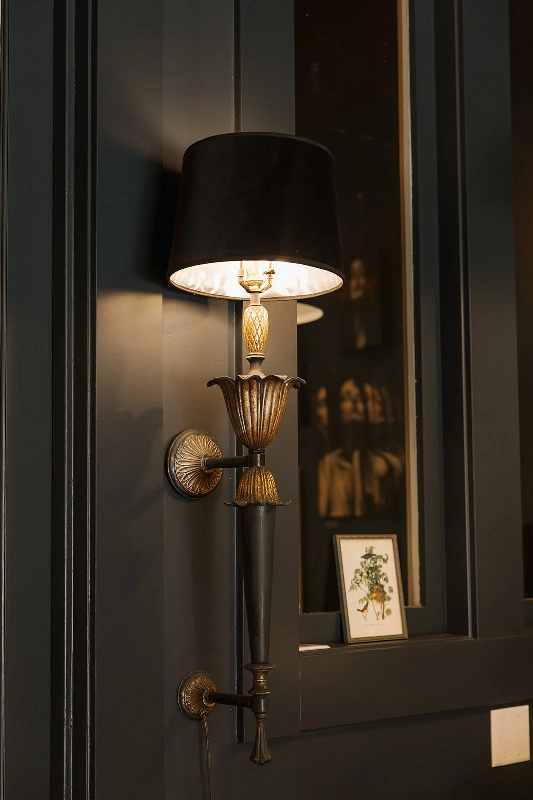 Decorative wall sconce with black lampshade, gold accents, mounted on a dark wall near a window with framed picture.