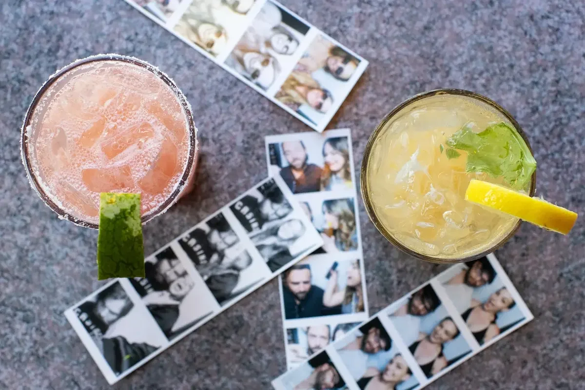 Two cocktails with lime garnishes on a table scattered with photo booth strips of various people.