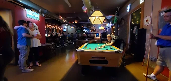 People playing pool at a bar with hanging lights and patrons seated at tables in the background.